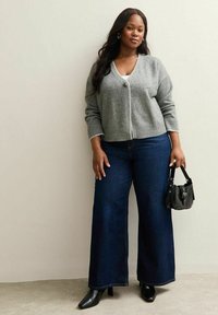 Cardigan grigio a maglia con scollatura a V e chiusura a bottoni, abbinato a jeans in denim blu scuro a gamba larga. Accessoriato con una piccola borsa nera.