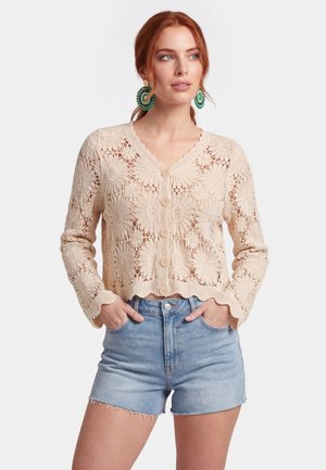 Vrouw met rood haar draagt grote groene oorbellen, een beige bloemige gehaakte cardigan die dichtgeknoopt is, en lichtblauwe denimshorts met handen in de zakken.