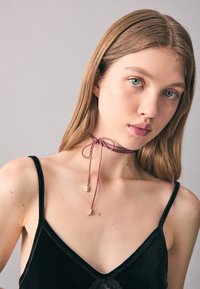 Na krku má obepínací náhrdelník z mauve stuhy s kovovými zlatými doplňky, svázaný v mašličku vpředu. Modelka má na sobě černý sametový kamisol.