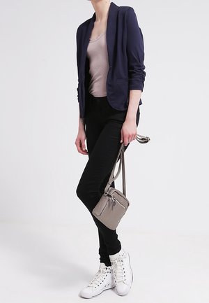 Marineblazer over een beige tanktop, zwarte skinny jeans en witte sneakers. Een grijze schoudertas met een ritsaccent. Eenvoudige, moderne outfit.