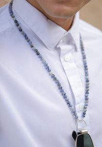 Collier en perles avec des pierres bleues et grises, présentant une boucle en métal argenté tenant des lunettes de soleil noires, porté sur une chemise blanche.