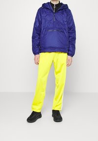 Blå, lätt pulloverjacka med huva och ficka med dragkedja fram, matchad med ljusgula träningsbyxor med sidelister och elastiska muddarna. Svarta sneakers.