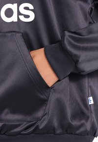 Svart satinjacka med en stor vit logotyp, ribbade ärmslut och en sidficka med en synlig blå märkesetikett.