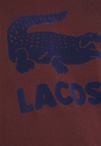 T-shirt bordeaux avec un logo crocodile bleu foncé et un texte audacieux lisant "LACOSTE." Tissu lisse avec un design classique à col rond.