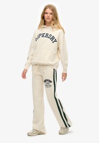 Kremowa bluza z kapturem z napisem "SUPERDRY" w granatowym, łukowatym napiszie, w połączeniu z kremowymi spodniami z ciemnozielonym paskiem i logo. Miękki, luźny krój.