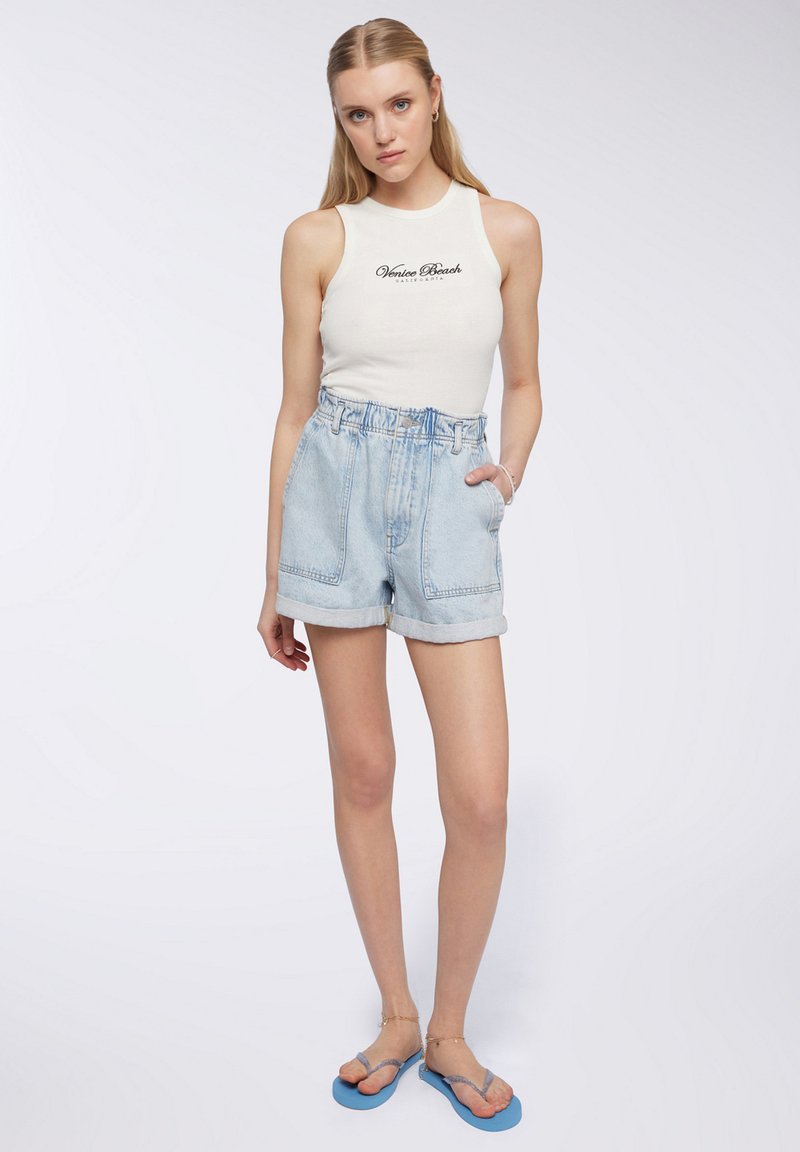 Giovane donna con lunghi capelli biondi, indossa un top bianco senza maniche con la scritta "Venice Beach", shorts di jeans azzurri e infradito blu.