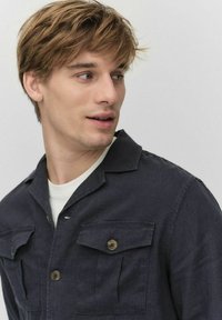 Donkerblauwe button-up shirt met twee borstzakken, gestructureerde stof en een kraag. Draagt boven een wit T-shirt. Model staat iets naar links gericht.