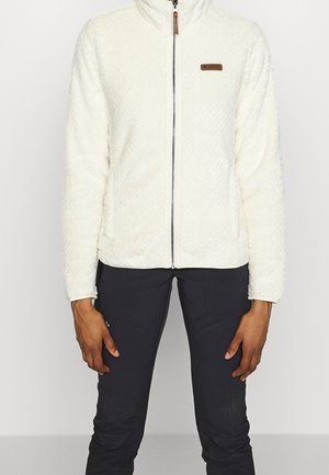 Persoon die een witte, getextureerde fleecejas met rits en zwarte broek draagt, staande tegen een effen lichte achtergrond.