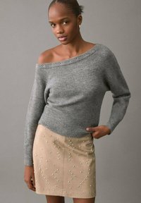 Sivi pleteni džemper s dizajnom koji otkriva ramena i metalnim detaljima s otvorima, uparен s bež suknjom s ukrasnim perlicama.