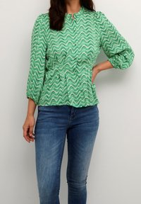 Donna che indossa una blusa verde a fantasia ondulata con bottoni e maniche a 3/4, abbinata a jeans aderenti blu, mentre posa con una mano sulla vita.