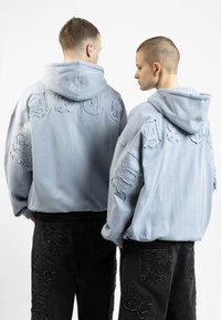 Des sweatshirts à capuche surdimensionnés bleu clair présentent des détails usés et des accents brodés sur le dos. Un jean sombre avec des patchs texturés complète le look.