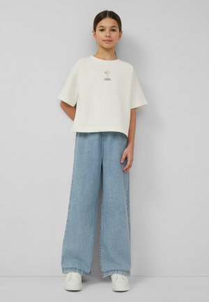 Dziewczyna stojąca przy szarej ścianie ubrana w białą koszulkę z krótkim rękawem z małym kwiatowym wzorem, szerokie jasnoniebieskie jeansy oraz białe trampki.