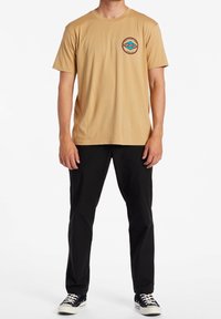 Camiseta de manga corta beige con un logotipo redondo que presenta olas azules y rojas en el lado izquierdo del pecho, combinada con pantalones negros y zapatillas.
