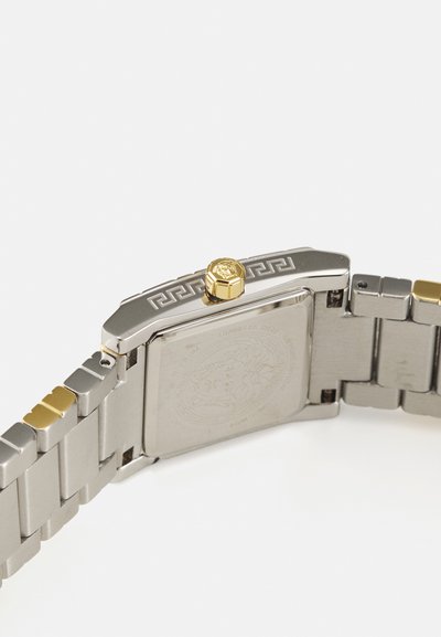 Hõbedane metallkell, millel on ristkülikukujuline korpus, kuldne kroon, mustriline raam ja vahelduvad hõbedased ja kuldsed lingid rihmal. Tekstuuriga tagakülg.