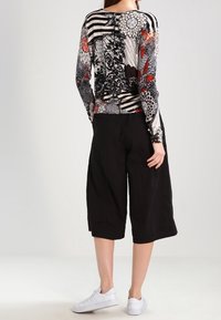 Kvinna som bär svarta vida cropped byxor, vita sneakers och en långärmad topp med svarta, vita och röda blommiga och randiga mönster, vänd bort.