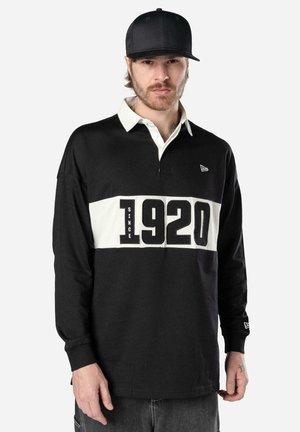 Mann mit schwarzer Baseballkappe und schwarzem Rugbyshirt mit weißem Kragen und dem Aufdruck "Since 1920" auf der Brust, steht vor einfarbigem Hintergrund.