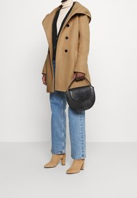 Kamel dobbeltbrystet frakke, sort struktureret håndtaske, beige turtleneck, tætsiddende sort vest, blå jeans og beige hælede ankelstøvler.