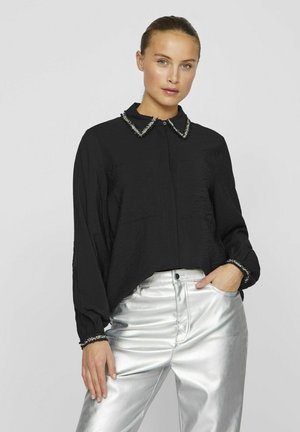Blusa nera con una finitura texturizzata, dotata di un colletto decorativo e chiusura con bottoni, abbinata a pantaloni argentati lucidi.