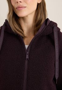 Sweat à capuche en polaire bordeaux avec fermeture éclair, tissu texturé et cordons de serrage, arborant un design imprimé "FAITES VOTRE PROPRE CHEMIN". Style à capuche.