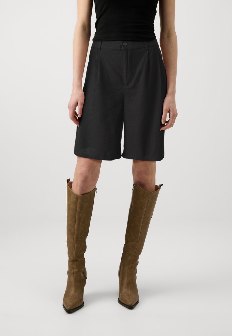 Femme portant un short noir jusqu'aux genoux, un haut sans manches noir, et des bottes marron montant jusqu'aux genoux avec des bouts pointus, debout devant un fond uni.