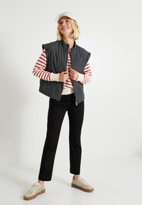 Gilet matelassé gris avec un col haut, porté sur un pull à rayures rouges et blanches, associé à un pantalon noir et des baskets beiges.