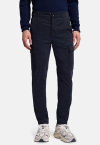 Pantaloni cargo navy in tessuto cotone, con tasche laterali cargo, chiusura a pulsante e vestibilità affusolata, abbinati a sneaker beige.