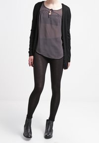 Svart cardigan över en genomskinlig grå topp med knyt-detalj, kombinerad med svarta leggings och ankelskor. Slät textur och figurnära design.