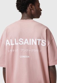Camiseta de gran tamaño en color rosa claro, hecha de algodón, con un audaz texto en blanco "ALLSAINTS" y caracteres japoneses en la parte trasera, con cuello redondo.