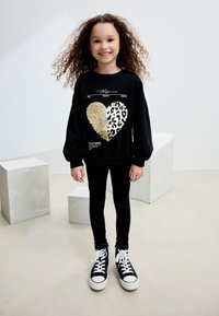 Felpa nera con grafica a cuore in stampa oro e leopardata, abbinata a leggings neri e sneakers alte. Capelli chiari e ricci.