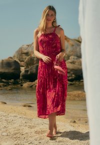 Maxi-Kleid in leuchtendem Pink mit einem Blumenmuster, Bandträgern und einem gestuften Design. Hält eine kleine pinke Handtasche an einem sandigen Strand.