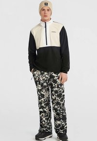 Mustast ja kreemvalgest fliispulloverist ees lükkekinnitusega, millele on kombineeritud mustad püksid valge lillemustriga. Aksessuaarideks beež nokamüts.