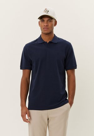 Navyblaues Polohemd mit kurzen Ärmeln, weicher Textur und Kragen. Mit subtilen Logodetails auf der Brust. Kombiniert mit hellen Hosen.