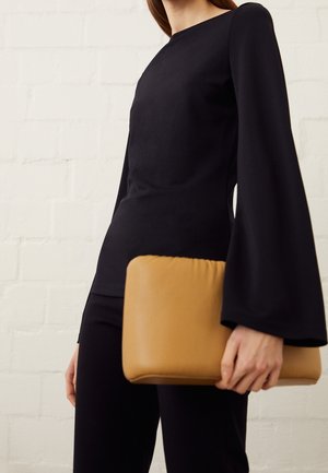 Personne portant un haut noir à manches longues larges, tenant une grande pochette rectangulaire couleur beige contre un mur de briques blanches.