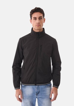 Uomo che indossa una giacca nera con cerniera, dotata di tasche sul petto e sui lati con zip, abbinata a jeans azzurri, rivolto verso l'obiettivo su uno sfondo neutro.