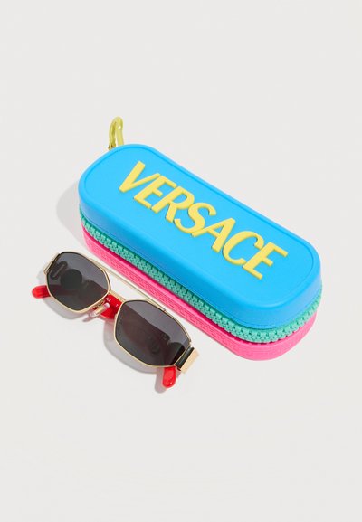 Μπλε, ροζ και πράσινη θήκη γυαλιών ηλίου Versace δίπλα σε γυαλιά ηλίου με χρυσή κορνίζα, μαύρους φακούς και κόκκινες άκρες στα μπράτσα, σε λευκό φόντο.