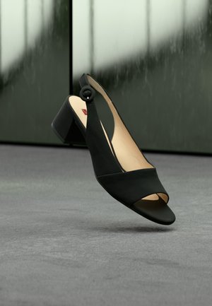 Μαύρο πέδιλο slingback με ανοιχτή μύτη και χοντρό τακούνι, τοποθετημένο πάνω σε γκρι επιφάνεια με θολό σκούρο πράσινο φόντο.