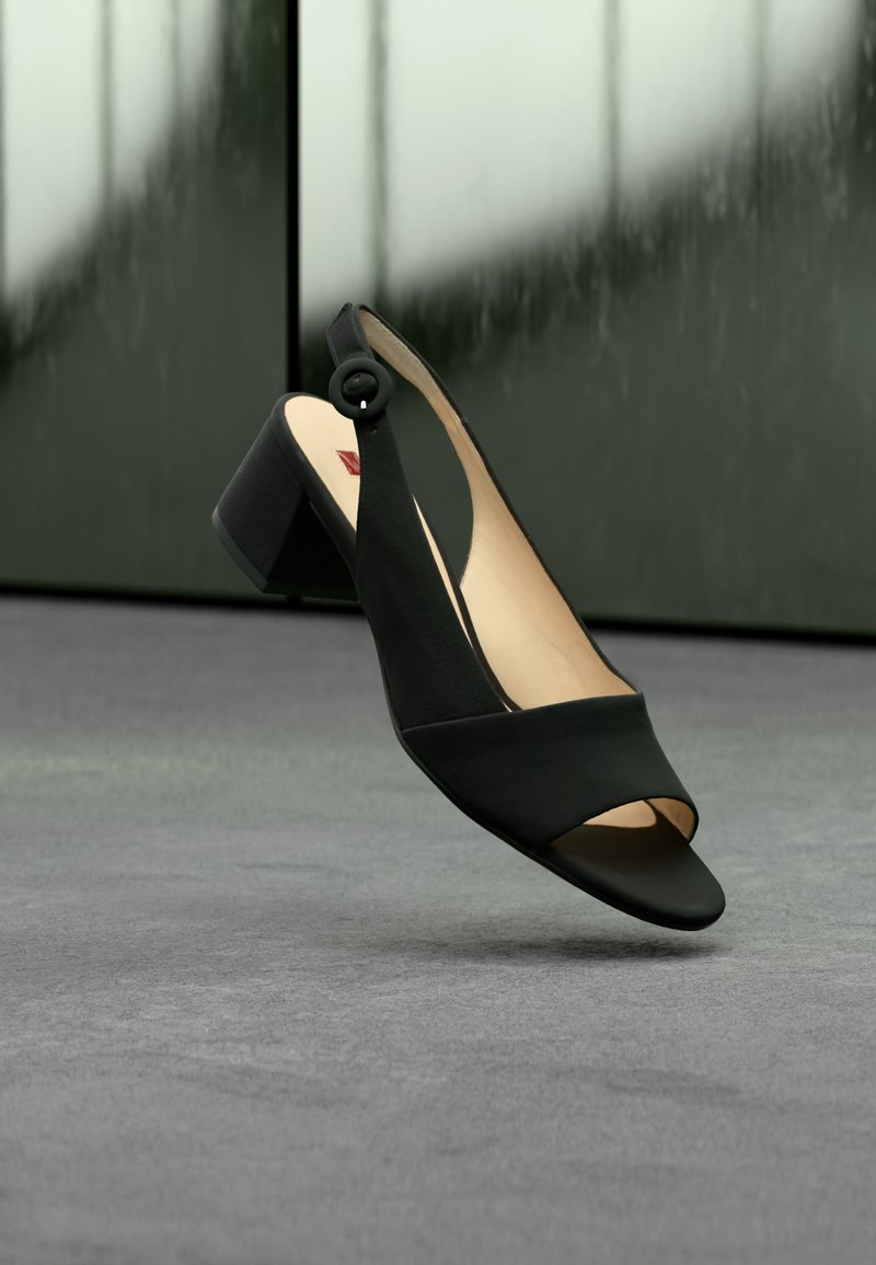 Μαύρο πέδιλο slingback με ανοιχτή μύτη και χοντρό τακούνι, τοποθετημένο πάνω σε γκρι επιφάνεια με θολό σκούρο πράσινο φόντο.