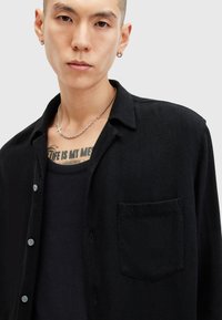 Chemise noire à boutons avec une finition texturée, poche poitrine simple et un col doux, superposée sur un débardeur noir.