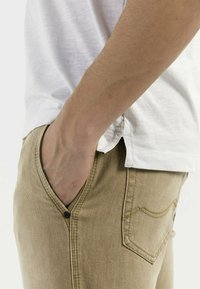 Beige denim broek met een voorzak en een knoopdetail. Gedragen met een witte katoenen t-shirt, waarbij een casual zijprofiel wordt getoond.