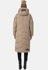 Long beige puffer coat with horizontal stitching, a large hood, side pockets, and a logo patch on the bottom left. Paired with black pants and boots.