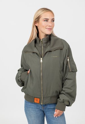 Vrouw in olijfgroene bomberjas met rits aan de voorkant, zijzakken en blauwe spijkerbroek, kijkt naar rechts met een lichte glimlach.