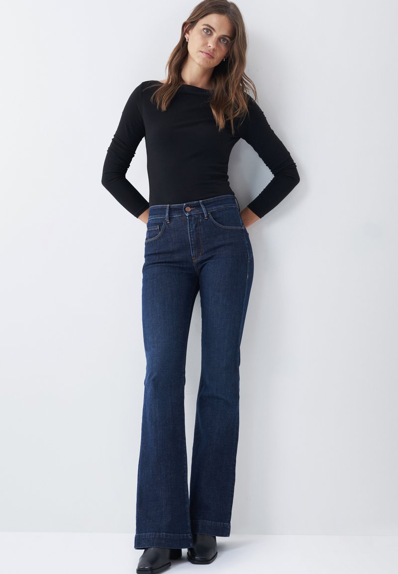 Jeans de mezclilla oscuros de talle alto y corte acampanado, combinados con una camiseta ajustada de manga larga negra. Los jeans tienen un clásico diseño de cinco bolsillos y cierre con botón.