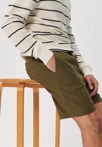 Khaki shorts with side pockets, made of cotton, paired with a white sweater featuring navy horizontal stripes. Wood stool beneath.