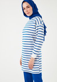 Vestido túnico a rayas en blanco con rayas azules, con mangas largas y aberturas laterales. Combinado con pantalones azules sólidos. Tejido suave, diseño casual.