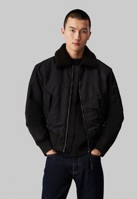 Chaqueta bomber negra con un grueso cuello de piel sintética marrón. Cuenta con un cierre frontal, bolsillos laterales y puños de canalé. Lleva puesta sobre un suéter oscuro.