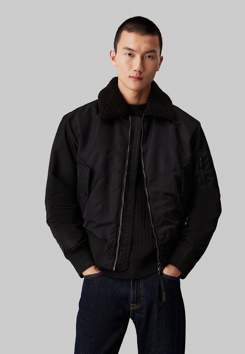 Chaqueta bomber negra con un grueso cuello de piel sintética marrón. Cuenta con un cierre frontal, bolsillos laterales y puños de canalé. Lleva puesta sobre un suéter oscuro.
