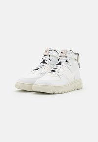 High-top sneakers in white leather with textured surface, cream rubber sole, black and tan accents, featuring laces and adjustable strap.
