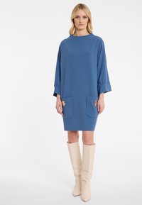 Eine Frau steht in einem blauen, lockeren Kleid mit langen Ärmeln, vorderen Taschen mit dekorativen goldenen Akzenten und kniehohen cremefarbenen Stiefeln.