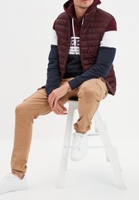Gilet trapuntato bordeaux sopra una felpa blu navy con strisce bianche e bordeaux, abbinato a pantaloni cargo beige e sneakers bianche, seduto su una scala bianca.