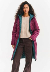 Dlouhý prošívaný kabát v barvě burgundska s tyrkysově modrým vnitřkem a kapucí. Spárováno s béžovým svetr s copánkovým vzorem a tmavě hnědými kalhotami.